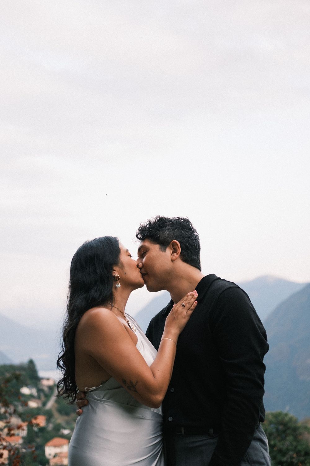 Couple sharing a romantic kiss outdoors with mountain landscape and cloudy sky in the background.
