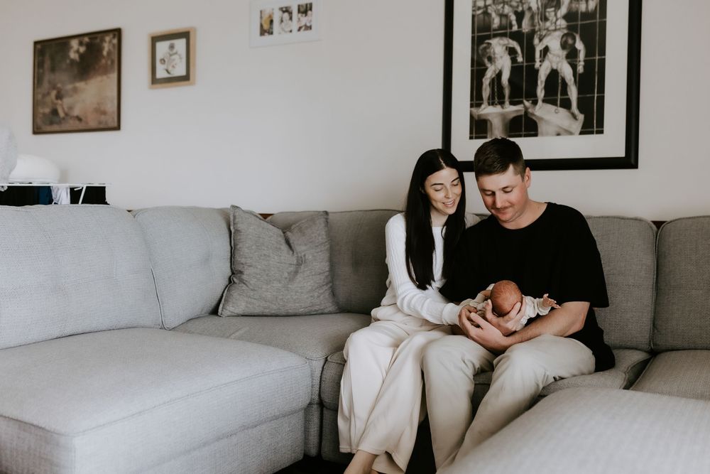 A couple sits on a gray sofa, lovingly gazing at their newborn baby in a cozy, modern living room.