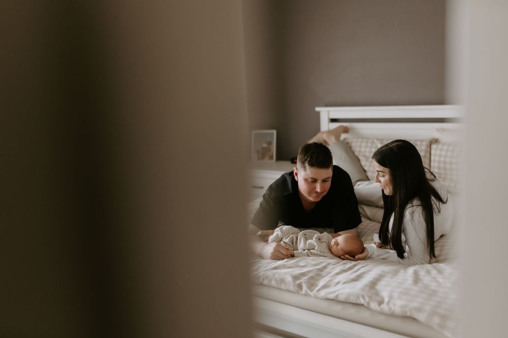 Parents lying on a white bed gazing lovingly at their newborn baby.