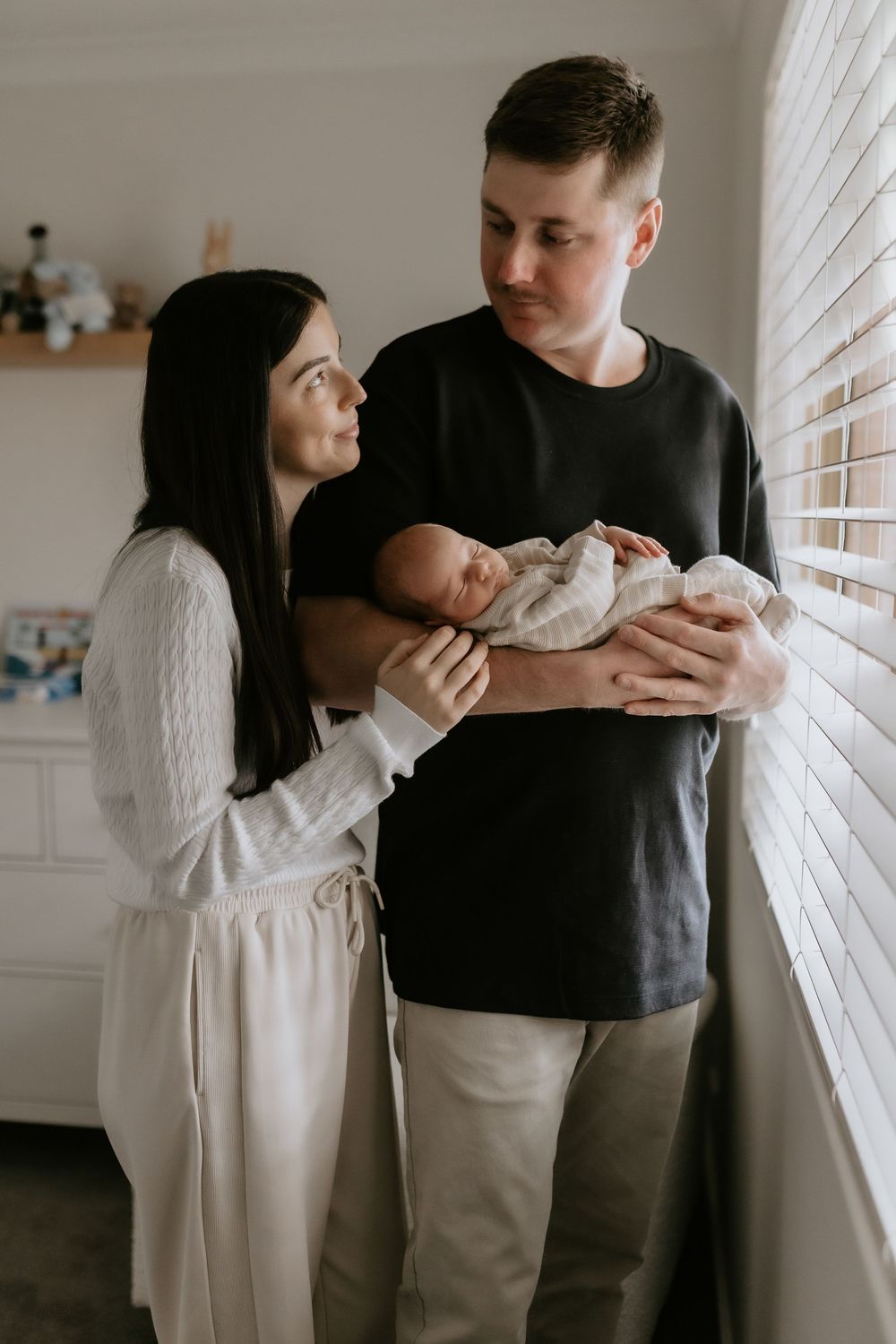 New parents lovingly gaze at their swaddled newborn baby by a window with soft natural light.