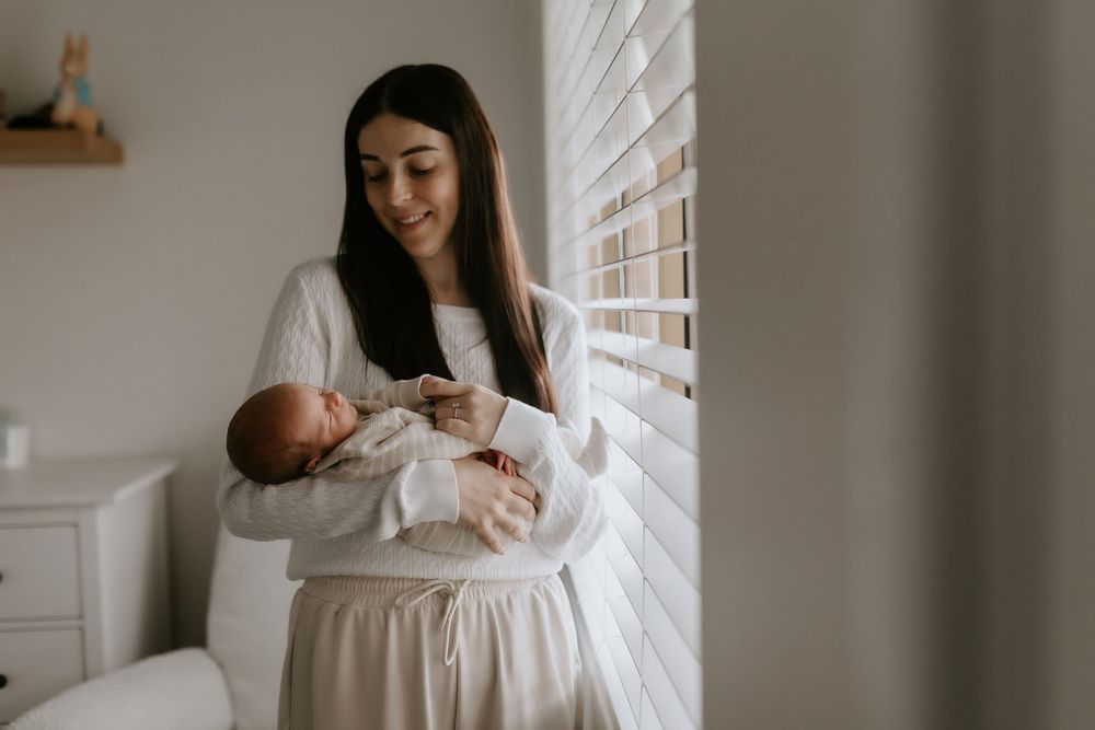 A smiling mother in white clothing holds her newborn baby near a bright window with white blinds.