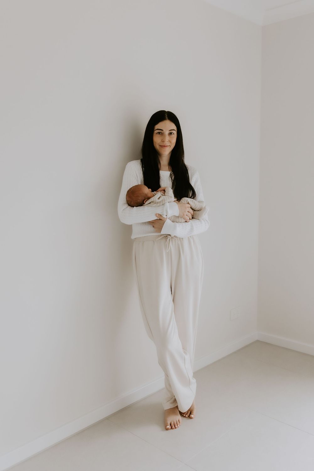 A woman with dark hair holds a newborn baby while leaning against a white wall in neutral loungewear.