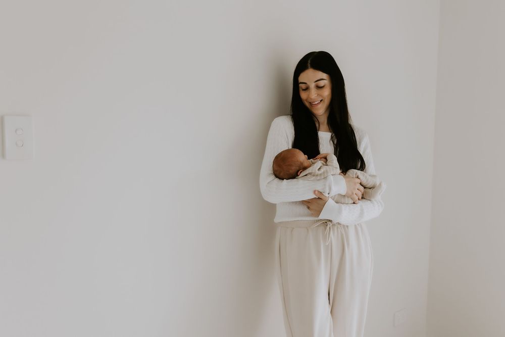 A smiling young mother with dark hair in white outfit tenderly holds her newborn baby against a light wall.