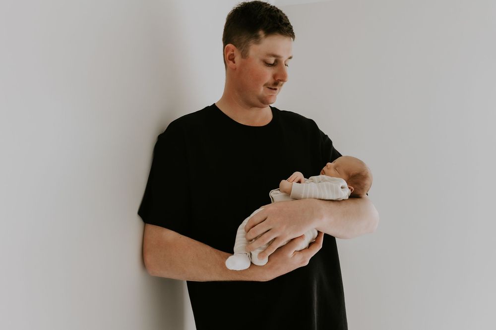 Man in black t-shirt tenderly holding a sleeping newborn baby against a white wall.