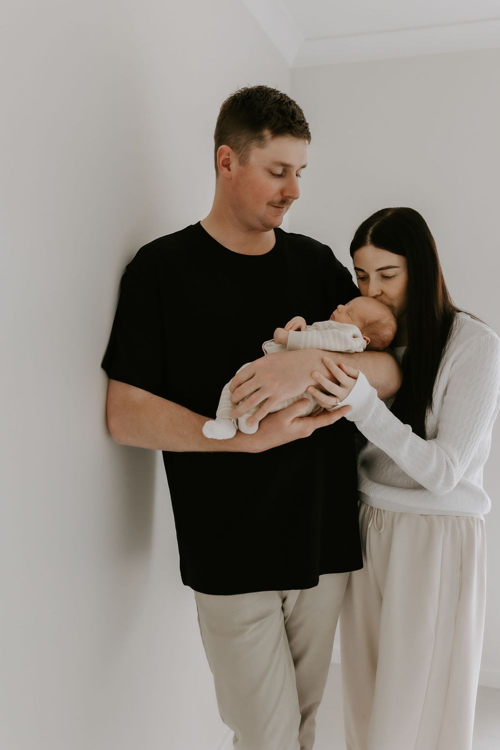 New parents tenderly hold their sleeping newborn baby, mother kissing infant as father looks on lovingly.