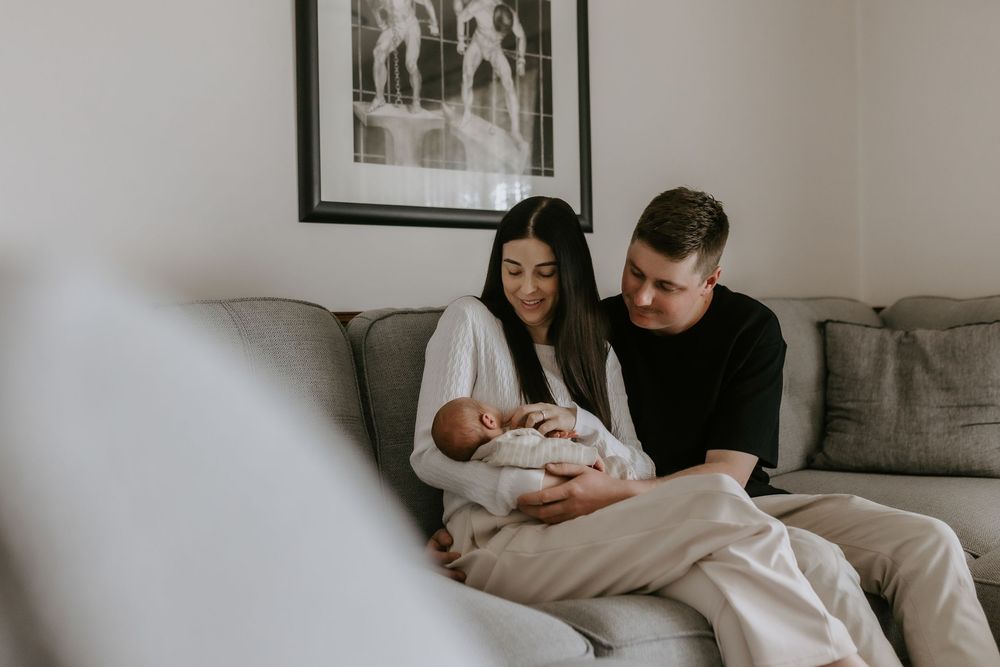A couple sits on a gray sofa, lovingly cradling their newborn baby in a cozy home setting.