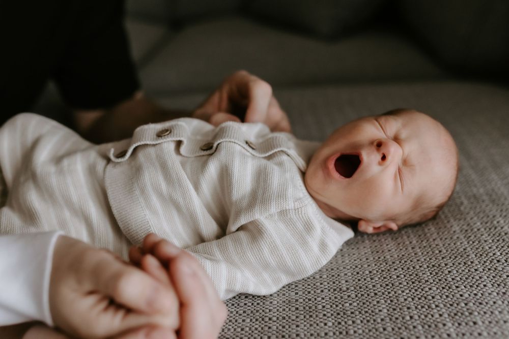 A newborn baby yawning while lying on a soft blanket, dressed in a cozy striped onesie, with an adult gently holding their hands.