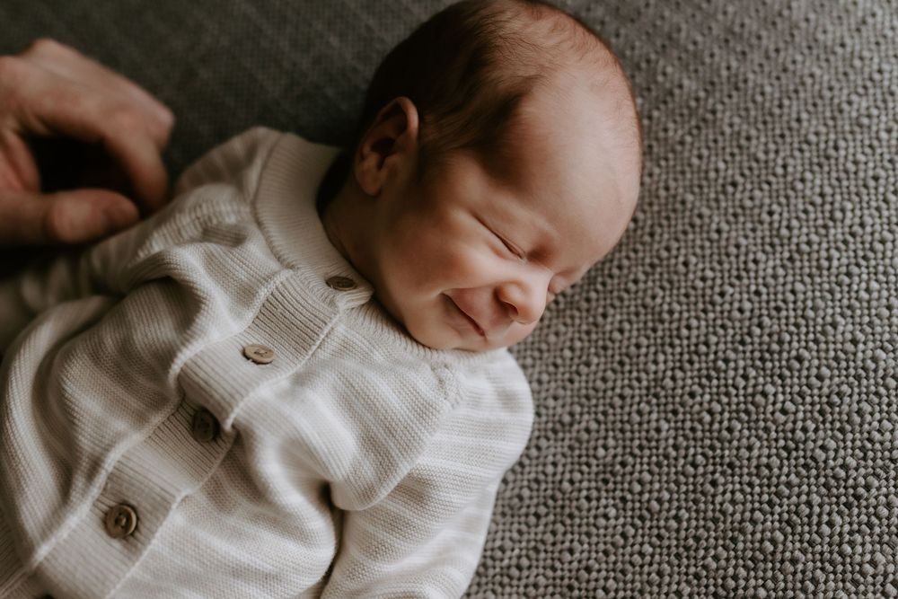 Peaceful newborn baby sleeping and smiling in a white knit onesie on a gray textured blanket.
