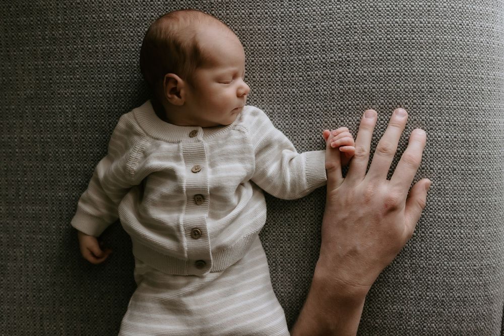 Newborn baby sleeping beside an adult hand on a gray textured blanket, wearing a striped onesie.