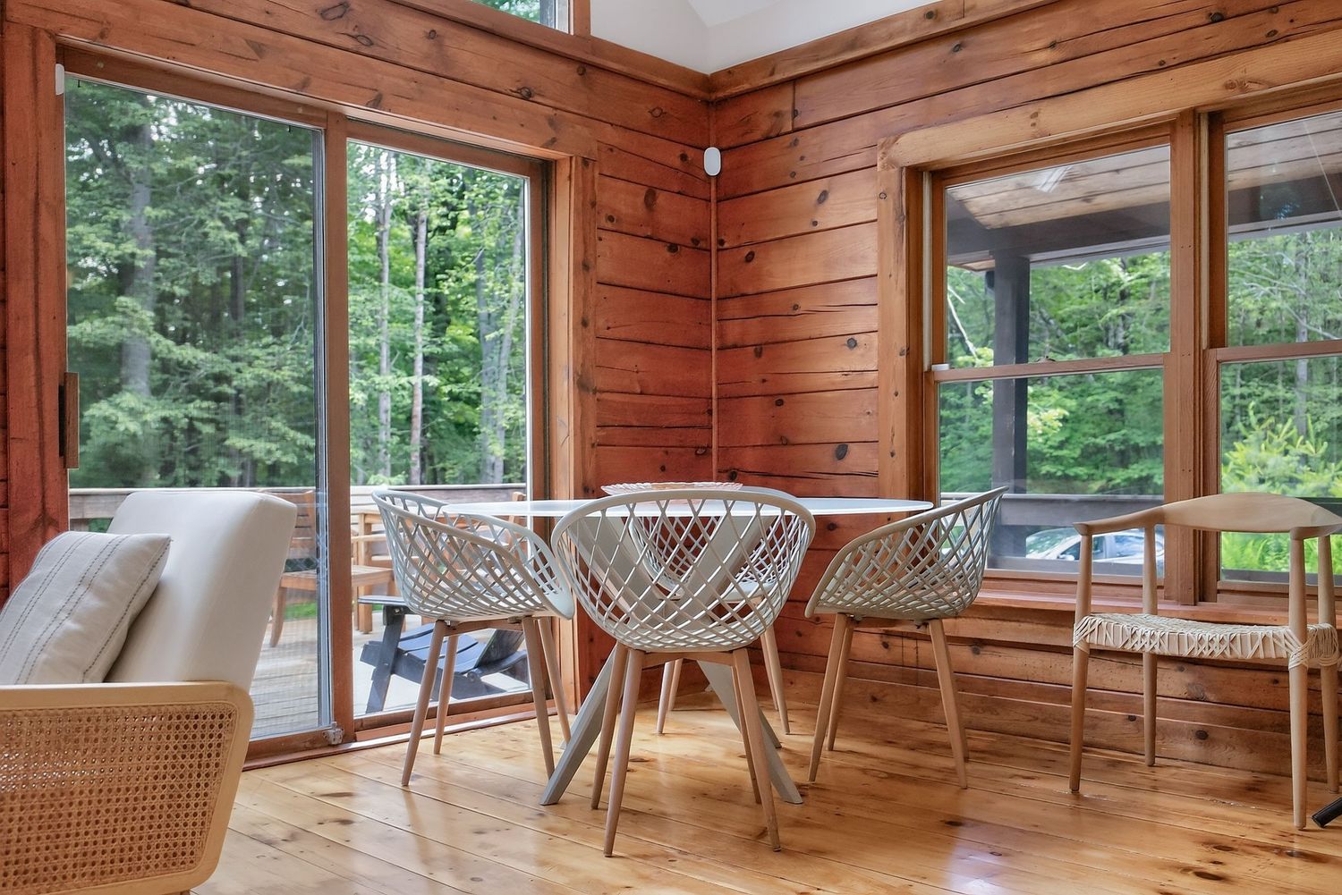 Bright sunlit log cabin dining area with white round table, wicker chairs, and large windows overlooking forest.
