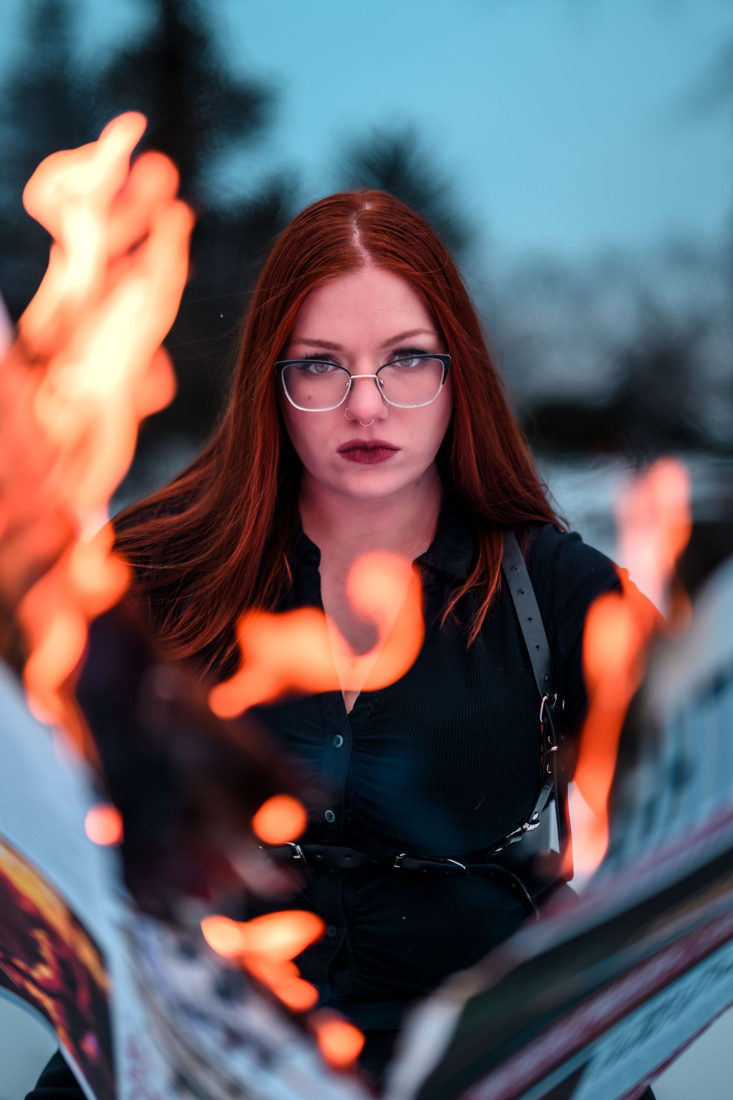 A young redhead woman with glasses in black outfit poses dramatically behind open flames in an outdoor setting.