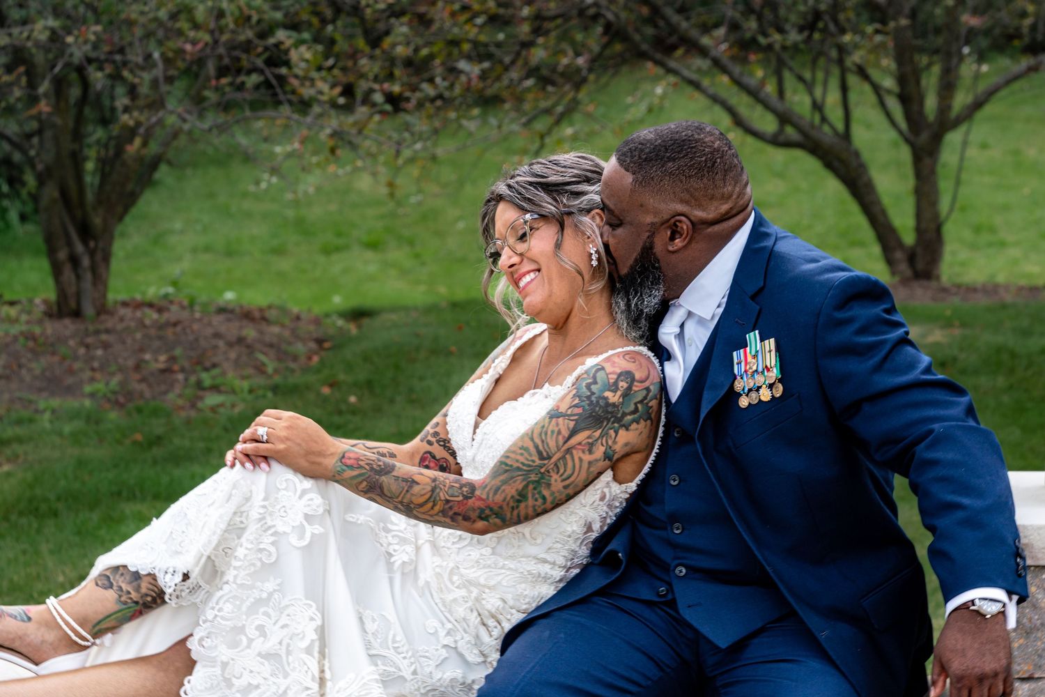 Bride in lace gown with tattoos sits with groom in navy suit wearing military medals outdoors.