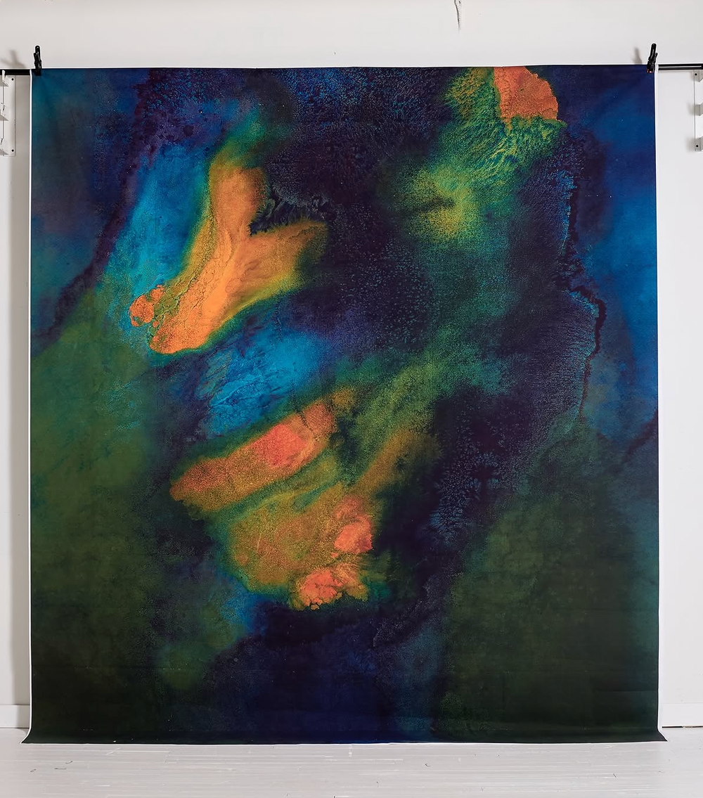 Abstract textile art featuring a face in vivid blue, green, and orange hues, hung on a white gallery wall.