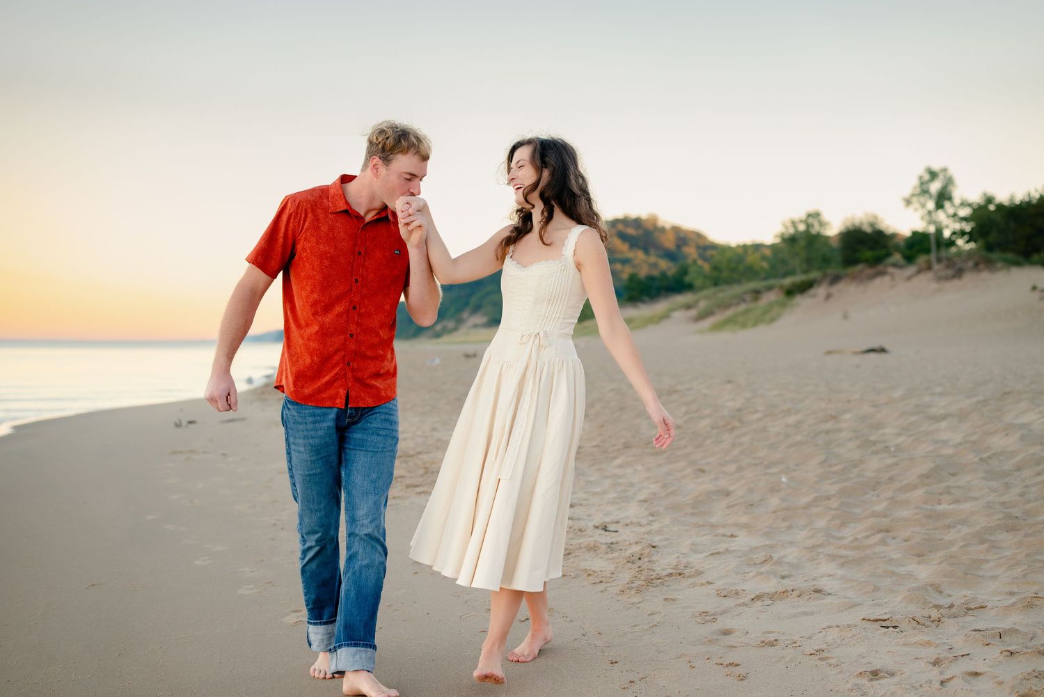 Couple walking barefoot on sandy beach at sunset, woman in white dress touching mans face, romantic seaside stroll.