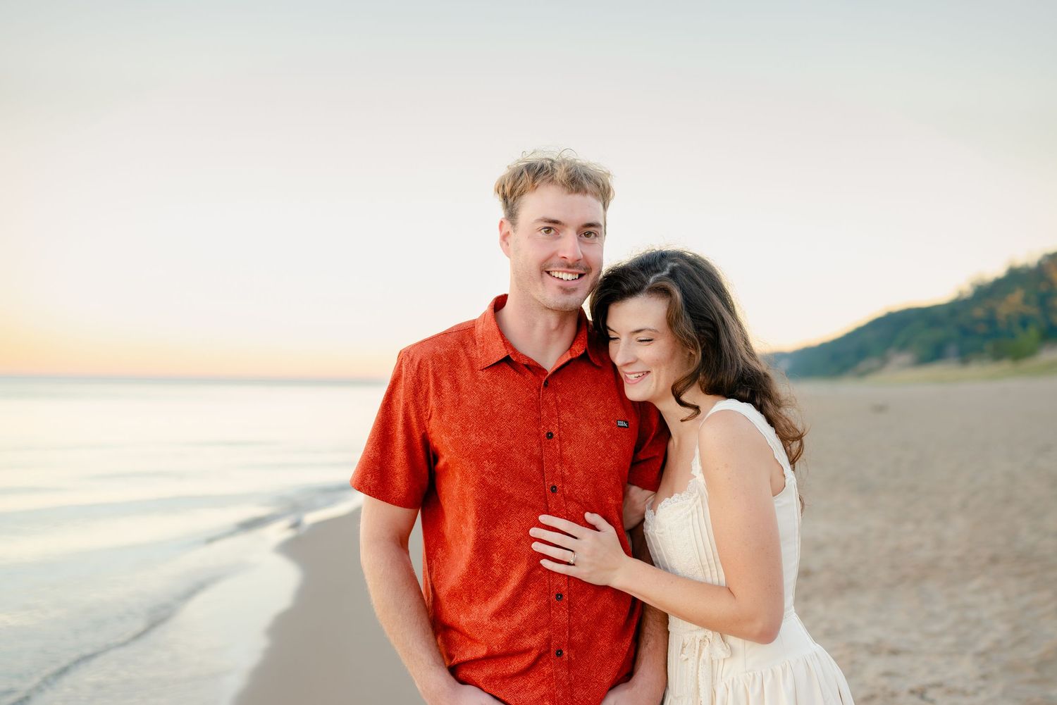 Couple smiling on a sandy beach at sunset, man in red shirt, woman in white dress leaning on his shoulder.