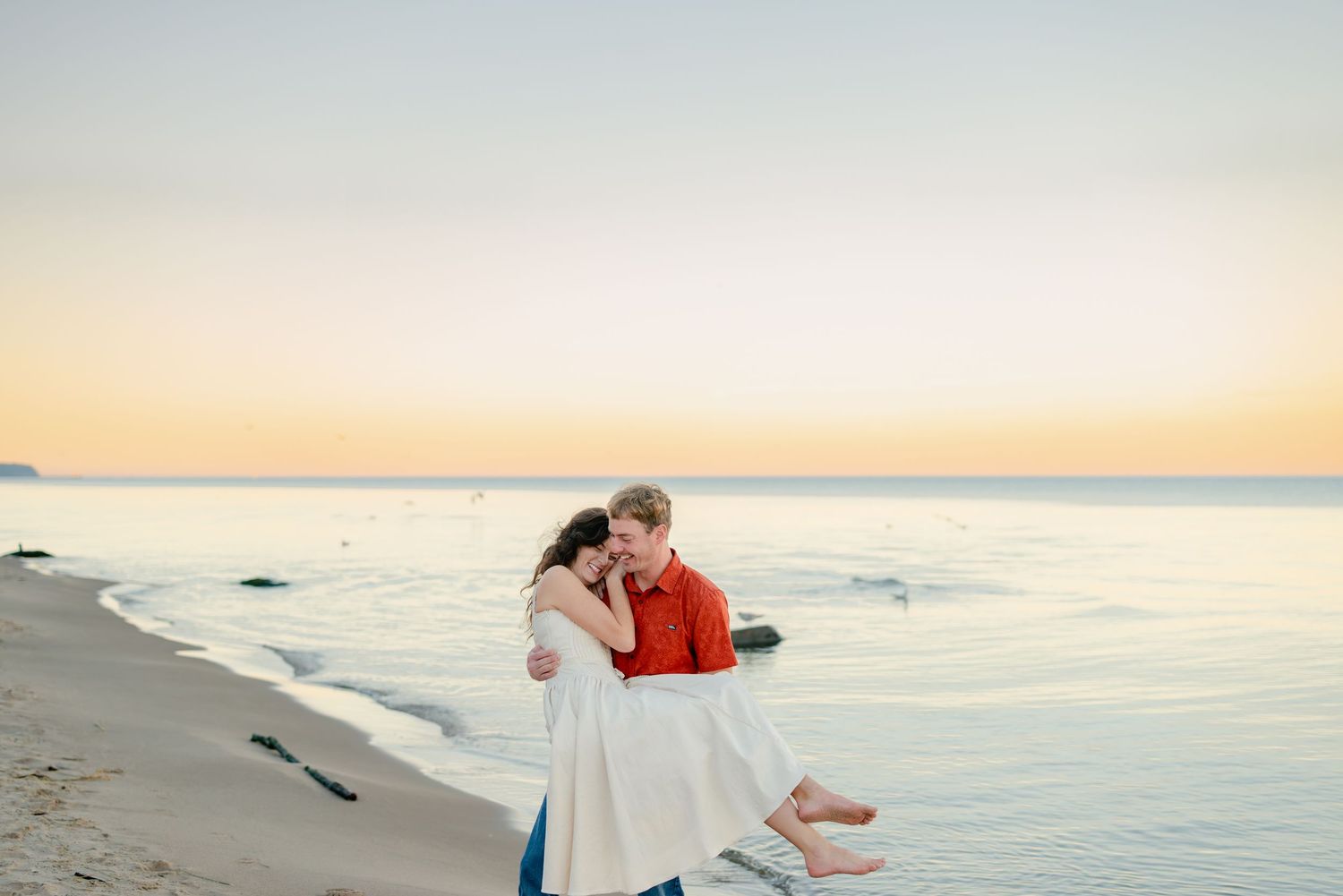 Couple sharing a romantic embrace on a sandy beach at sunset, man lifting woman in white skirt near calm ocean waters.