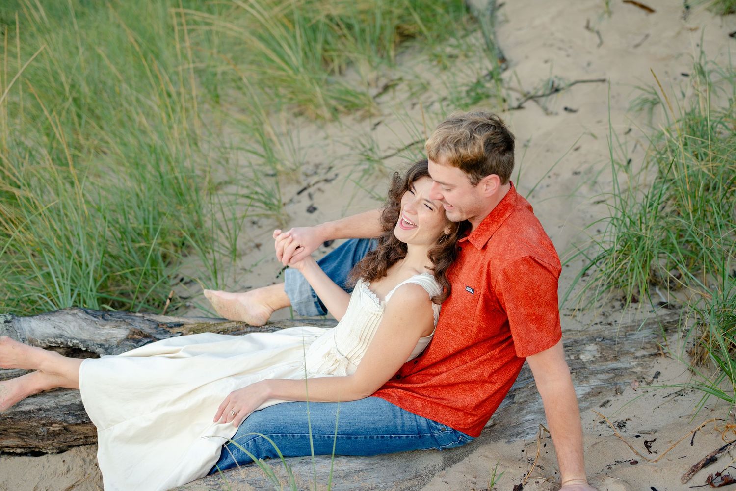 Couple sitting on driftwood in sandy dunes, woman in white dress leaning on man in red shirt, romantic outdoor portrait.