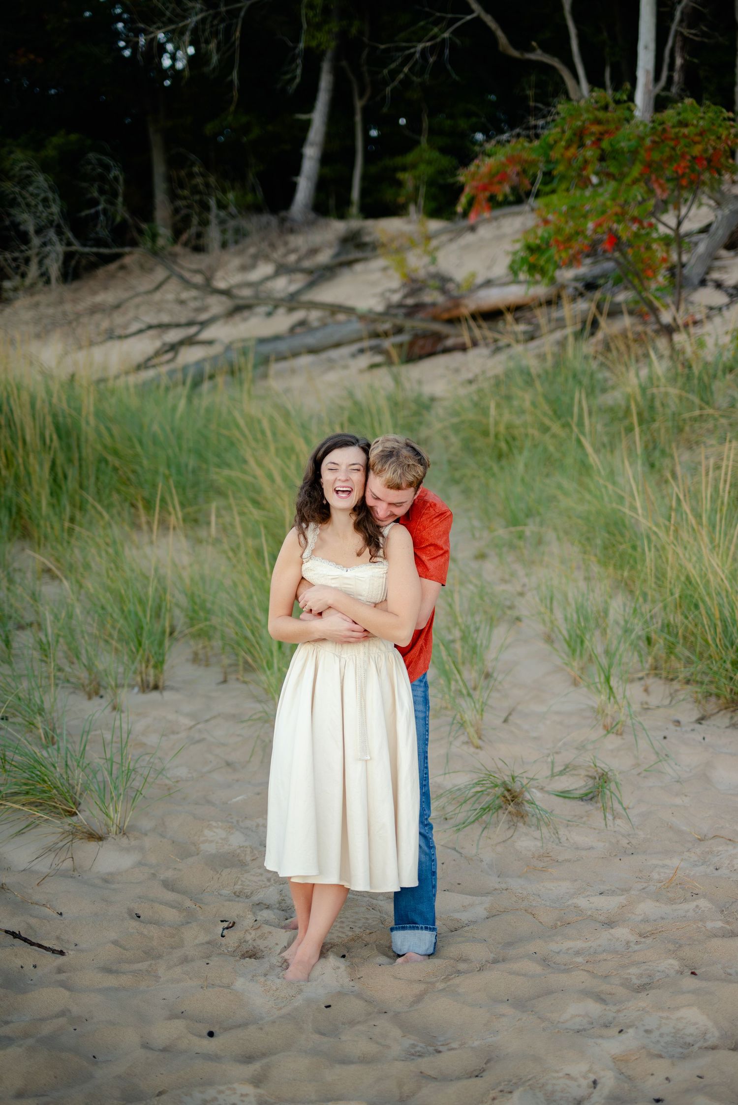 A couple embraces on a sandy beach, the woman in a white dress laughing joyfully as the man in a red shirt hugs her from behind.