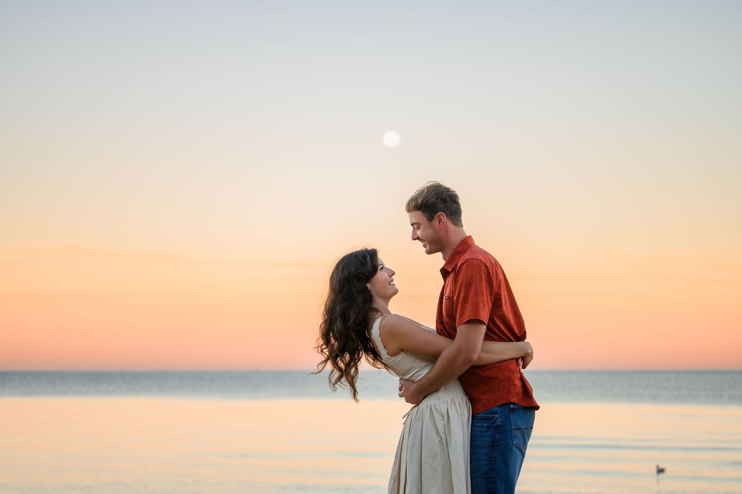 Couple embracing on beach at sunset with moon visible in pastel sky over calm ocean waters.