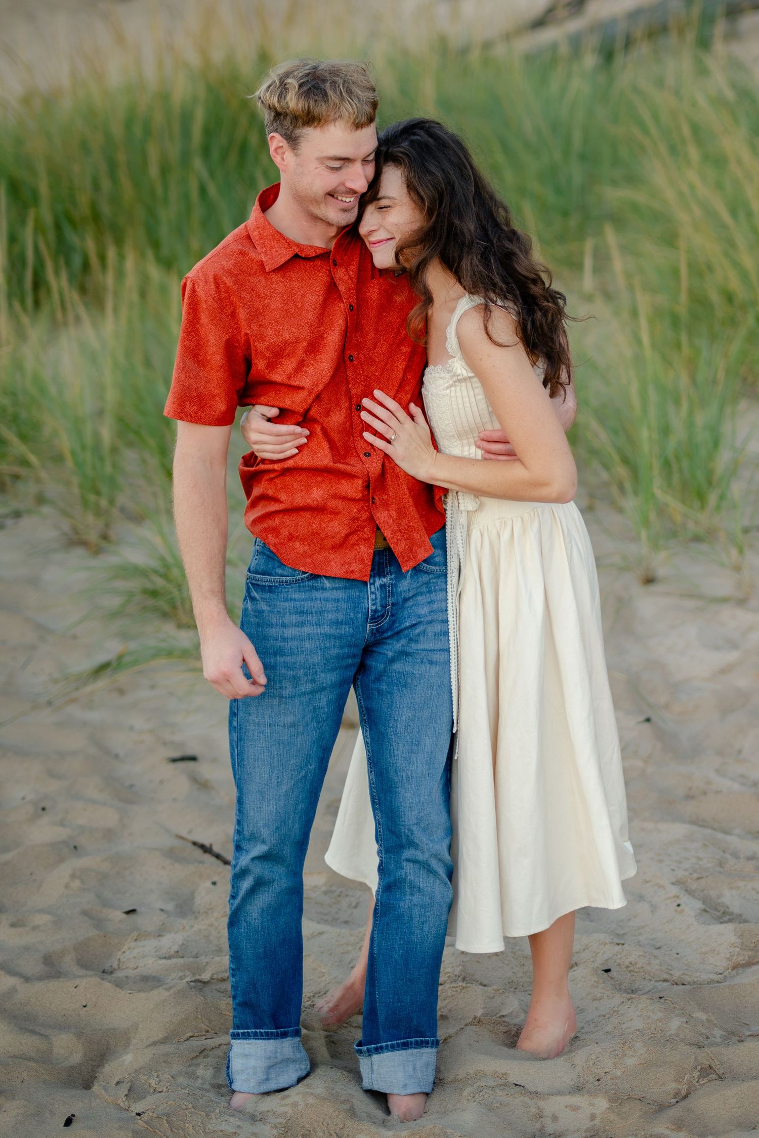 Couple embracing on sandy beach, man in red shirt and jeans, woman in cream dress, with green dunes behind.