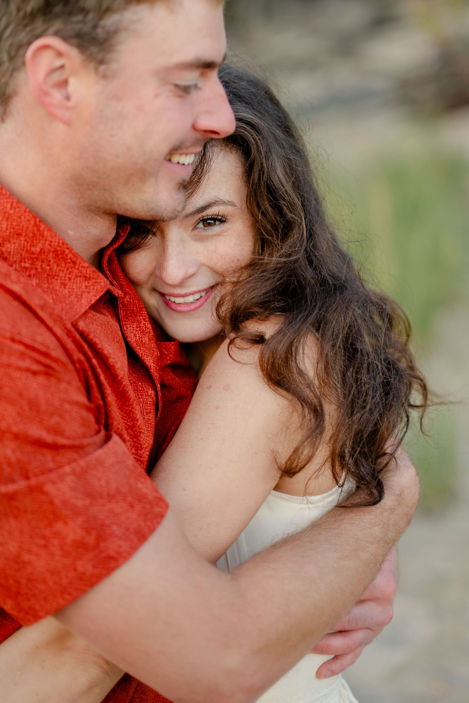 A smiling couple embraces outdoors, the man in a red shirt nuzzling the woman in a white dress.