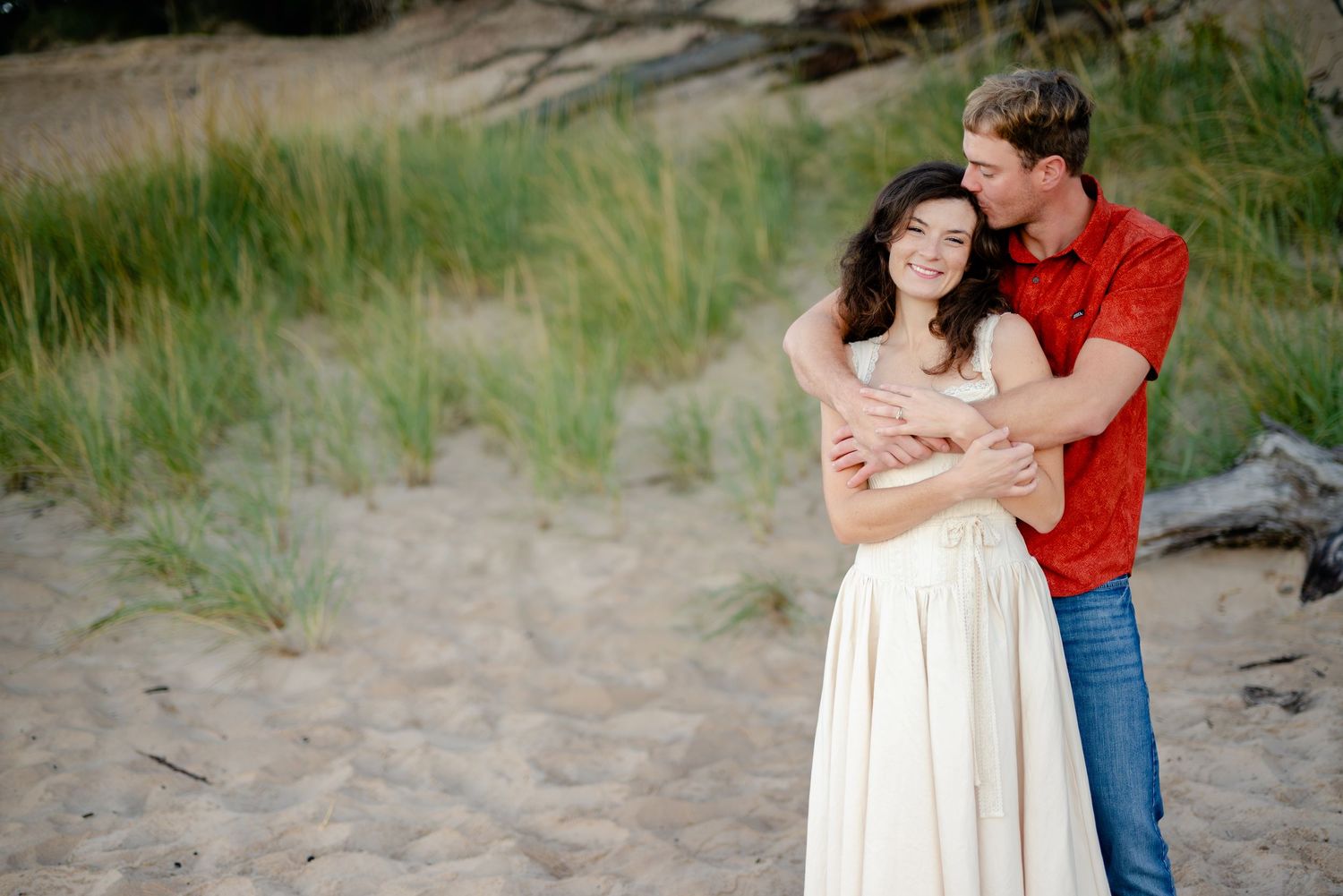 Couple embracing on sandy beach with green dunes, woman in white dress and man in red shirt sharing a romantic moment.