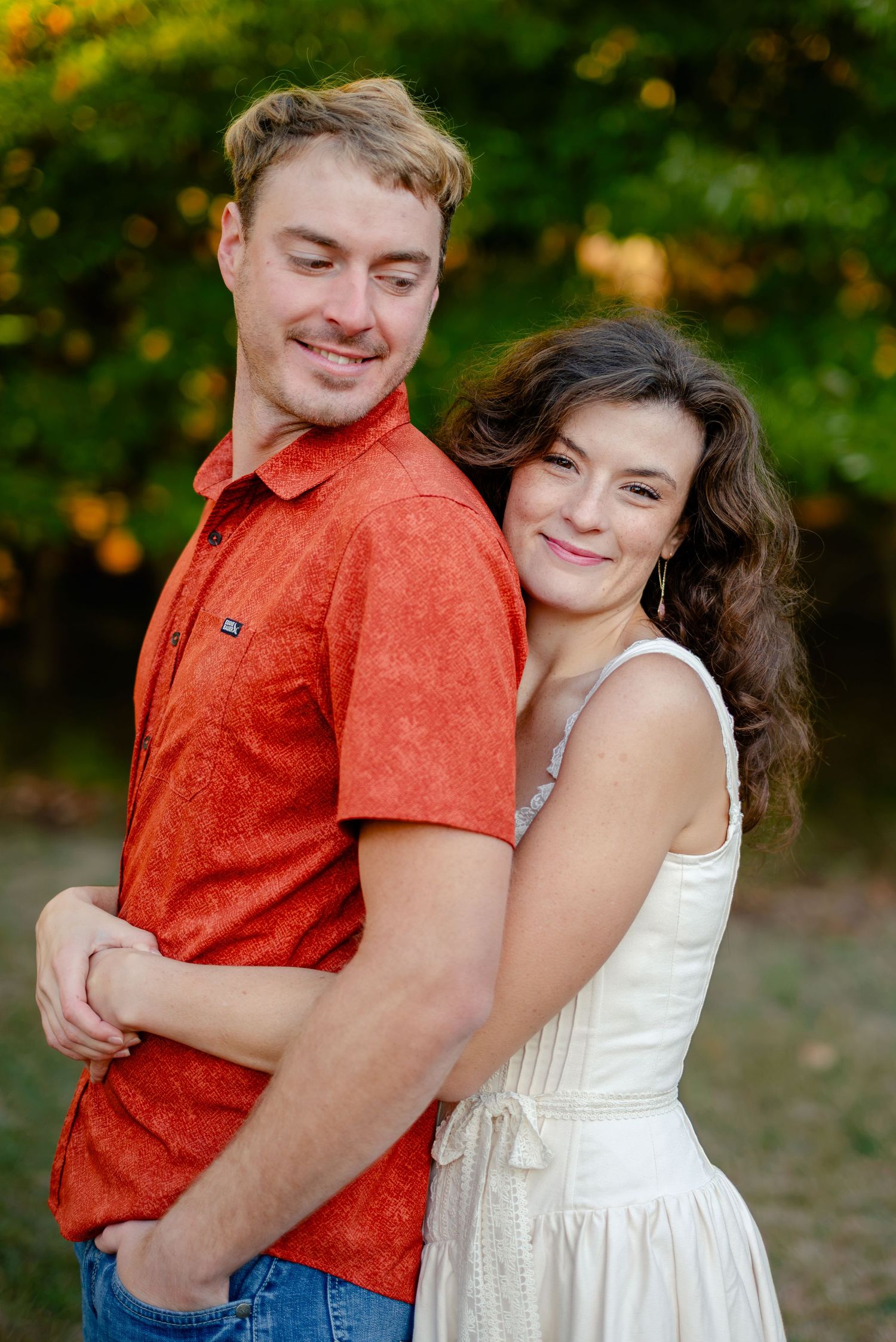 A smiling couple poses outdoors, man in red shirt and woman in white dress, embracing against a lush green background.