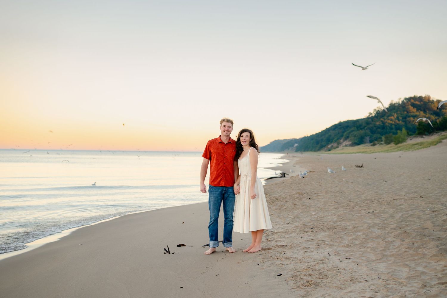 Couple posing together on a sandy beach at sunset with calm waves and a tree-covered hill in the background.