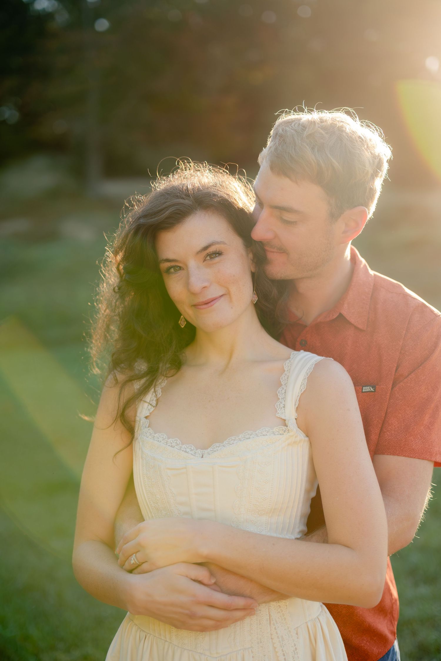 A couple embraces in golden sunset light, woman in cream dress with curly hair, man in red shirt kissing her cheek outdoors.