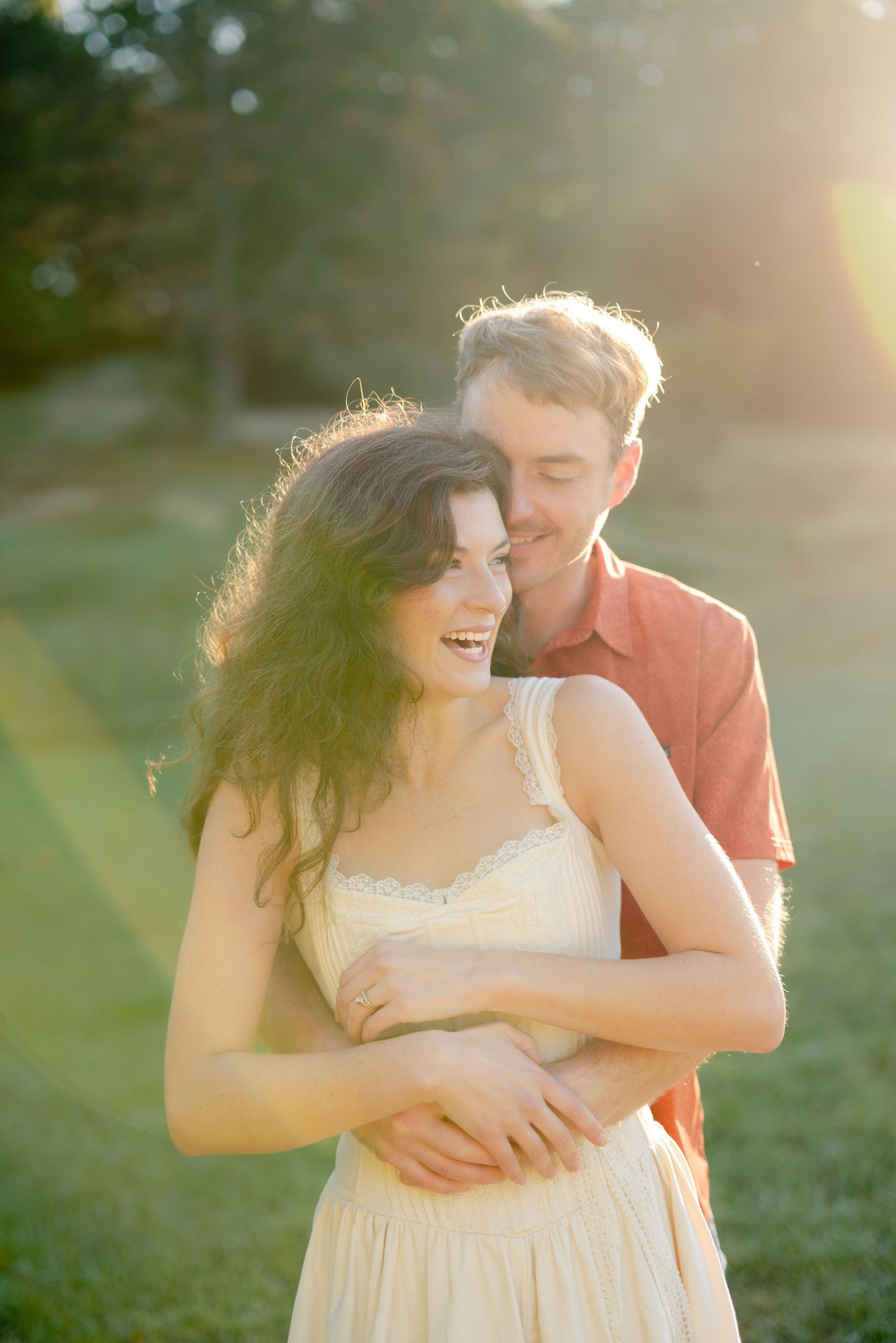 A couple embraces outdoors in golden sunset light, woman in cream dress smiling joyfully as man holds her from behind.