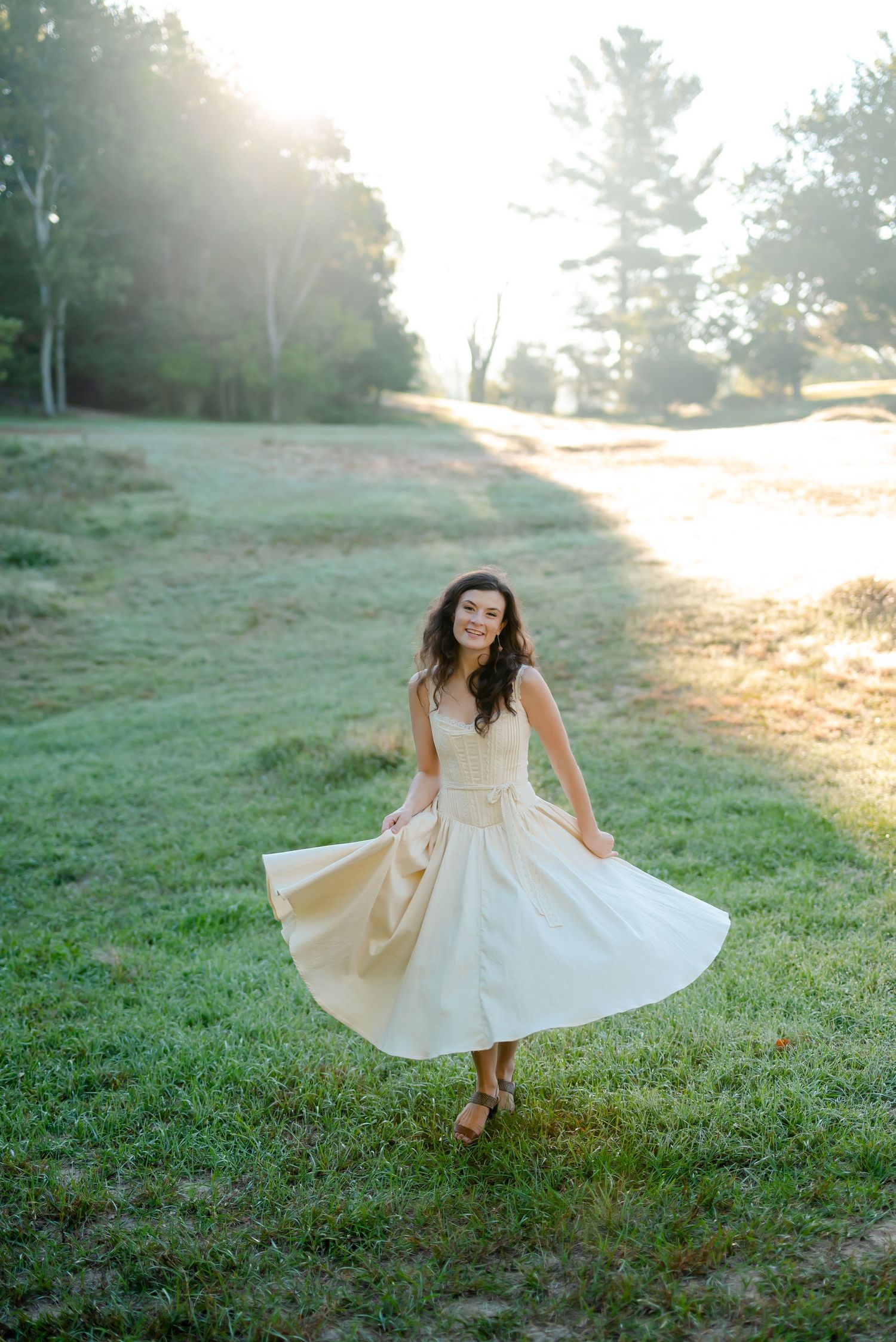 Young woman in a white midi dress twirling on green grass at golden hour with sunlight glowing behind trees.