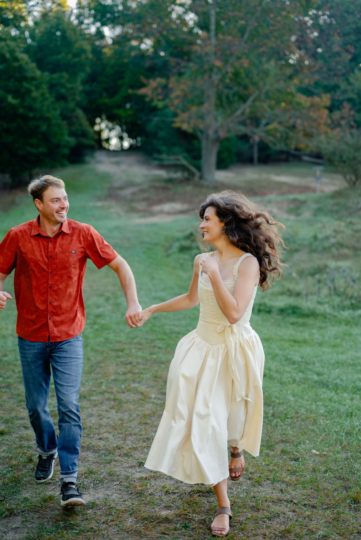 A joyful couple runs hand-in-hand through a lush green park, the woman in a yellow dress, the man in a red shirt.