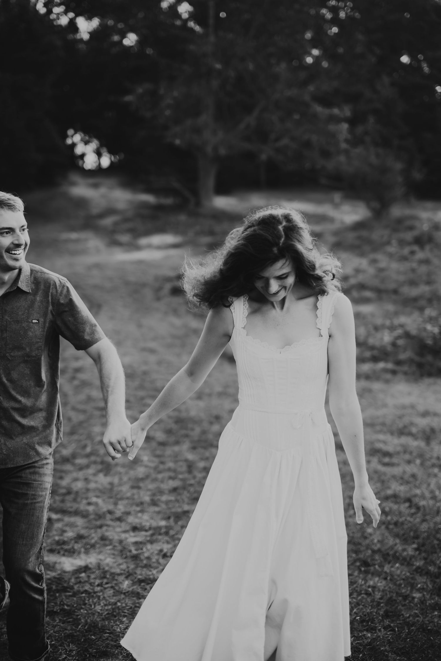 Black and white photo of a smiling couple holding hands while walking outdoors near trees.