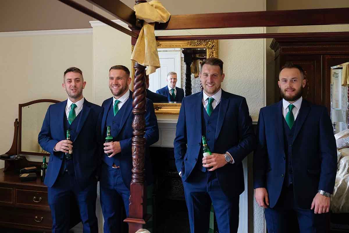 Four groomsmen in navy blue suits with green ties holding beer bottles posing together in a decorated room at Blaithwaite House