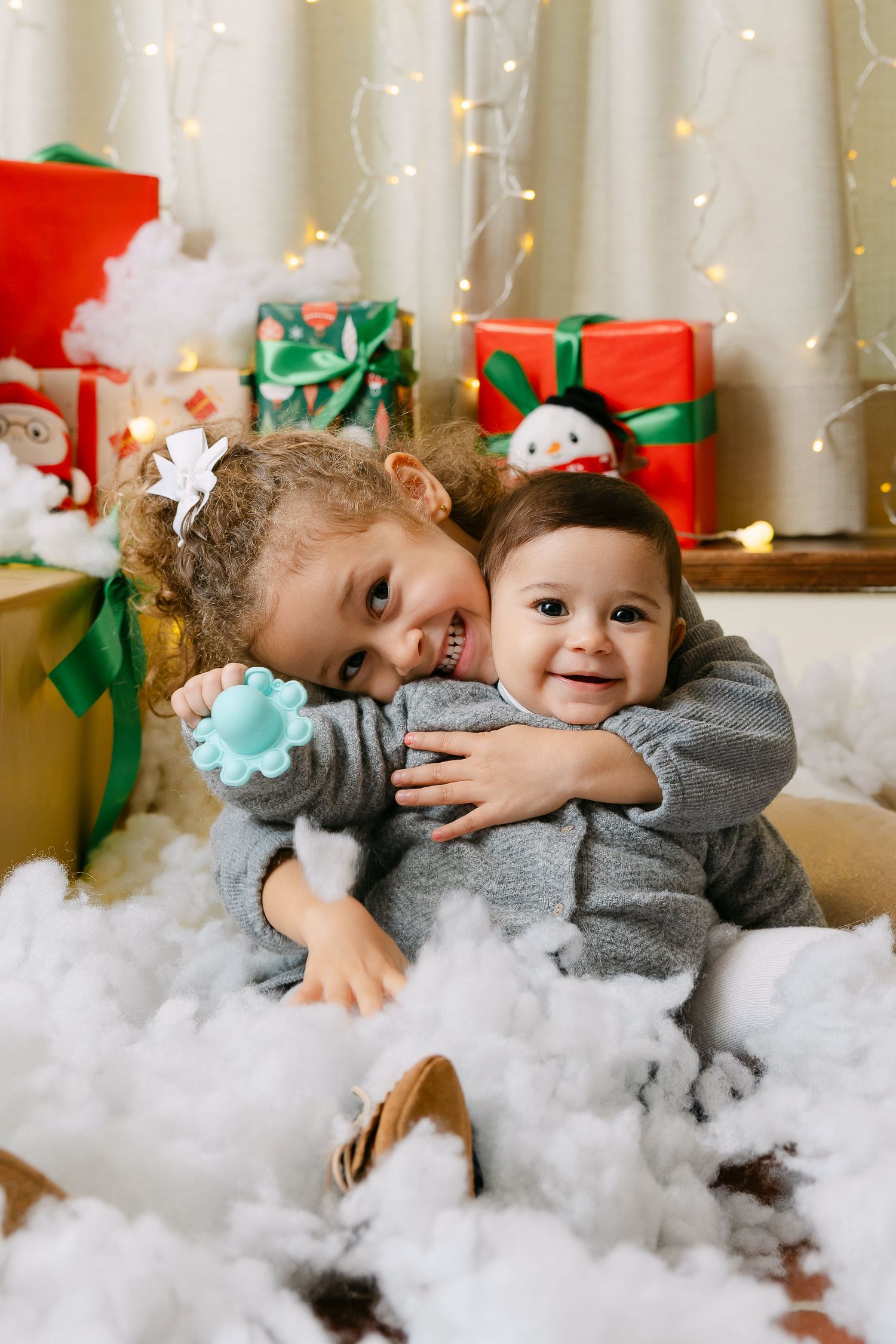 Two young children in gray outfits hug surrounded by Christmas gifts and lights in a festive holiday photo.
