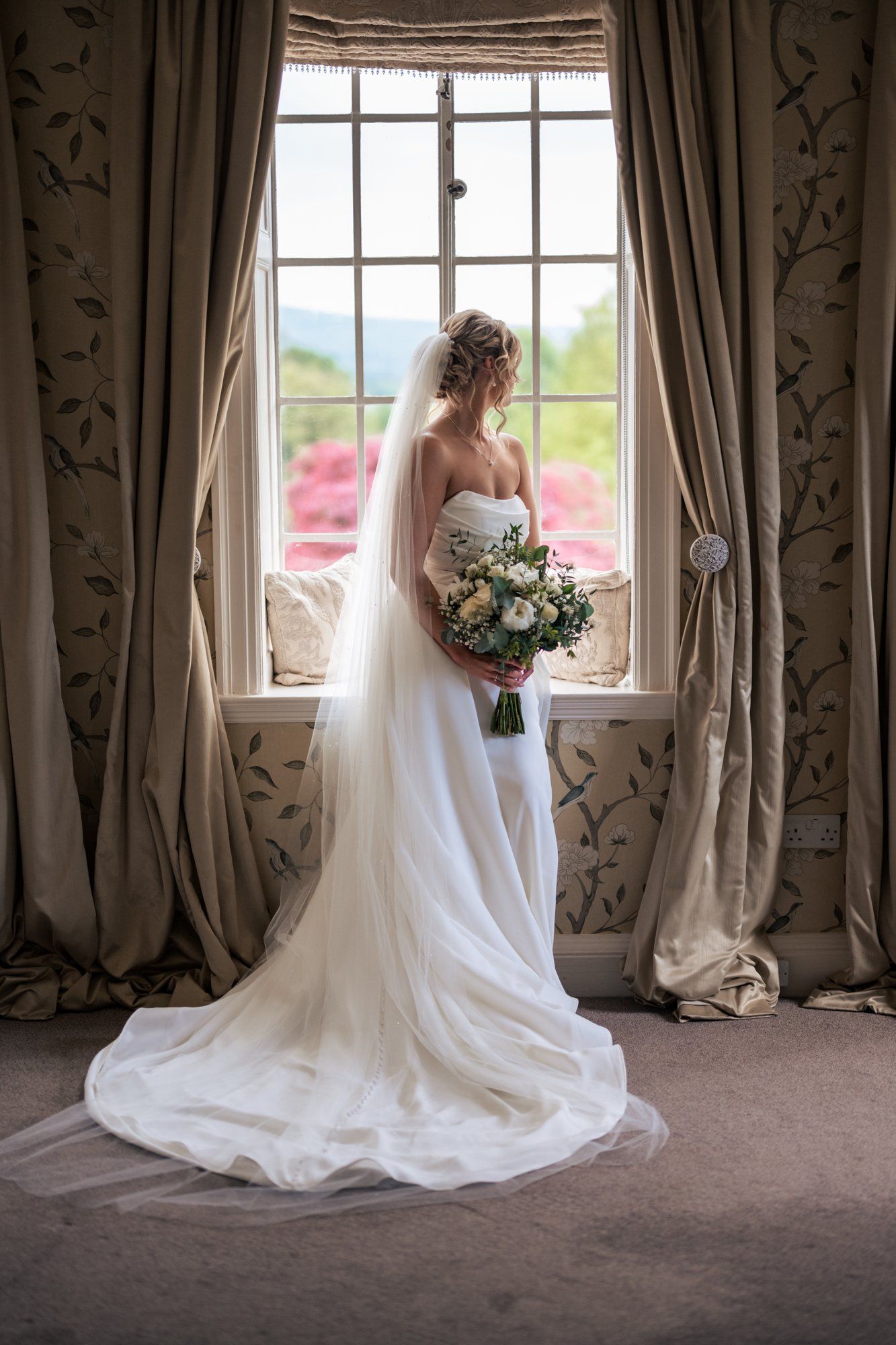 Bride in white gown and veil holds bouquet while standing before a large window with elegant drapes.