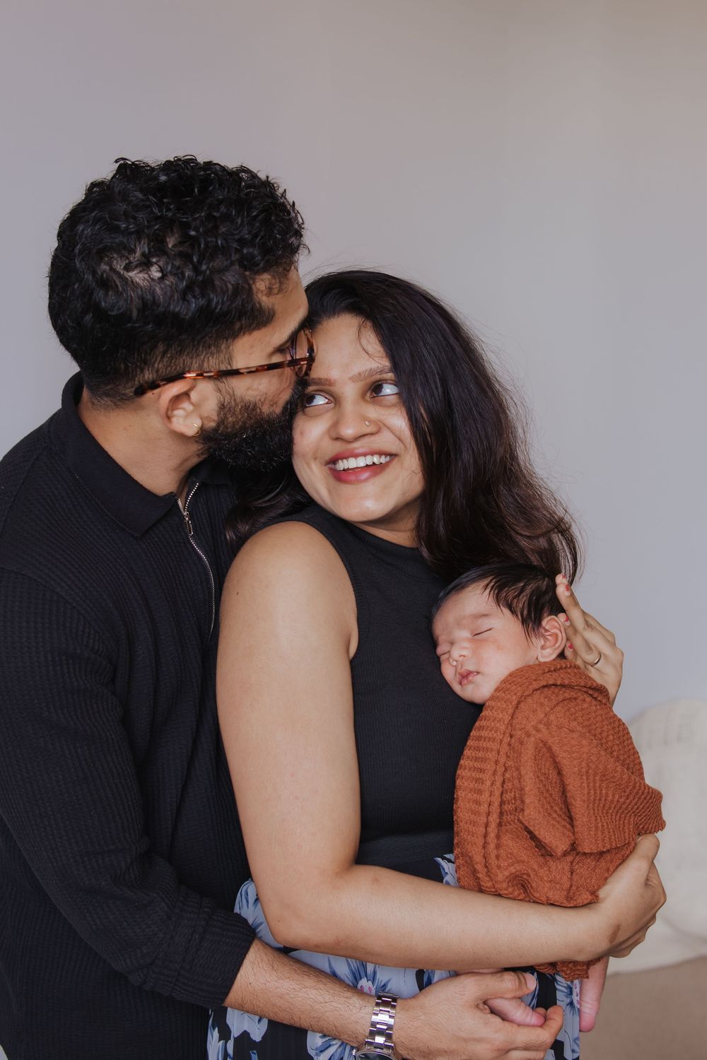 A couple smiles tenderly while holding their sleeping newborn baby dressed in a rust-colored knit sweater.