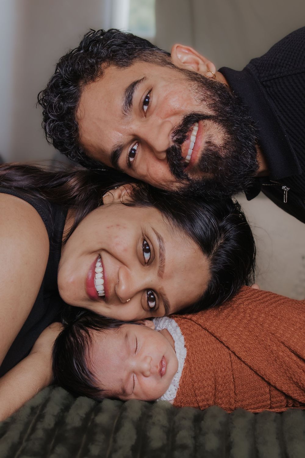 Happy family portrait with bearded father, smiling mother, and newborn baby lying together on a bed.