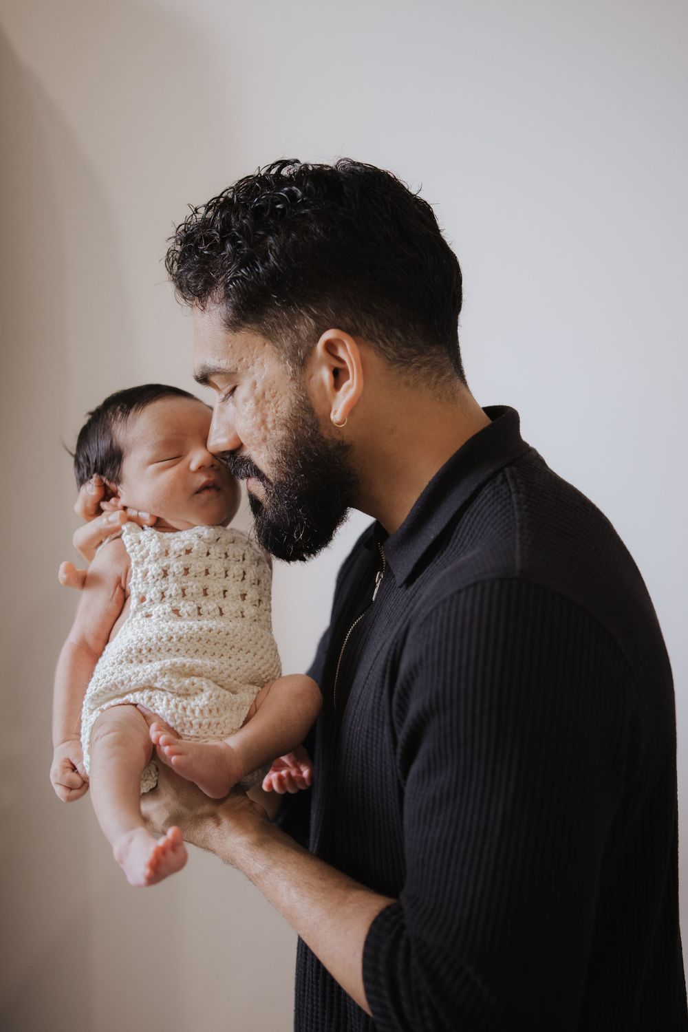 A bearded man in black gently kisses a newborn baby wearing a cream crochet outfit against a white background during in home newborn photos.