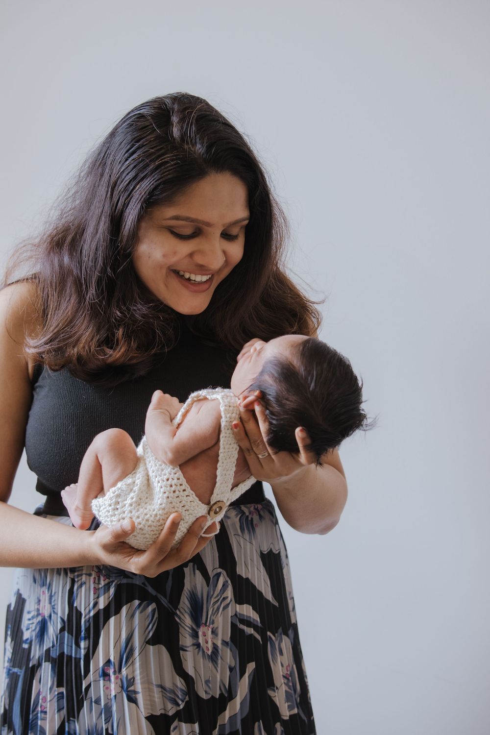 Smiling mother in floral skirt lovingly holds her newborn baby dressed in white crochet outfit.