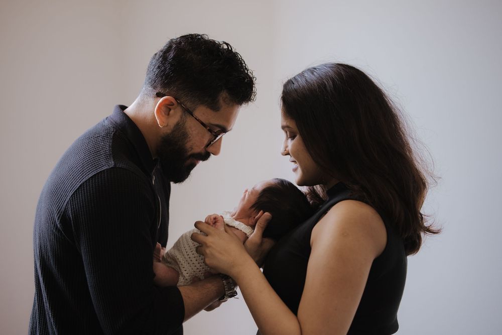 New parents in black outfits tenderly hold their newborn baby together against a soft white background.