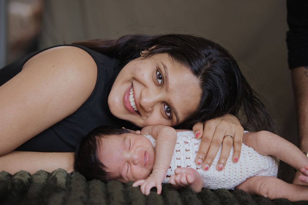 Smiling mother posing with her sleeping newborn baby wrapped in a white crochet blanket on a green surface.
