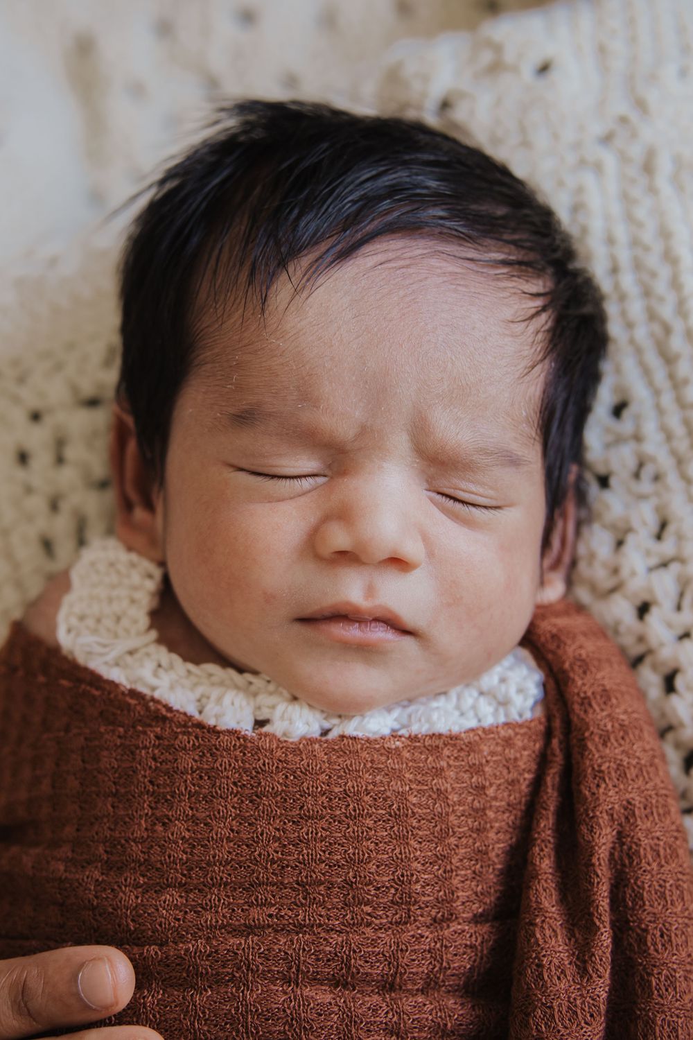 Peaceful newborn baby sleeping swaddled in a rust-brown knit wrap on a cream crochet blanket.