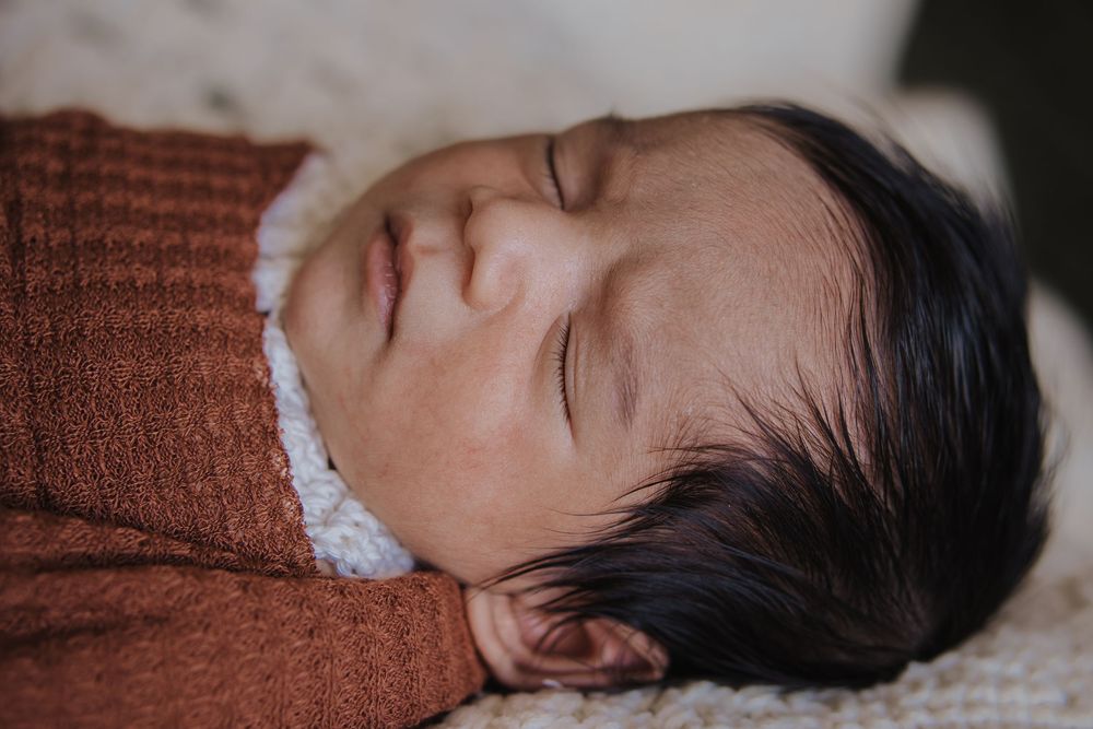Peaceful newborn baby sleeping soundly, wrapped in a rust-colored knit blanket with dark hair visible.