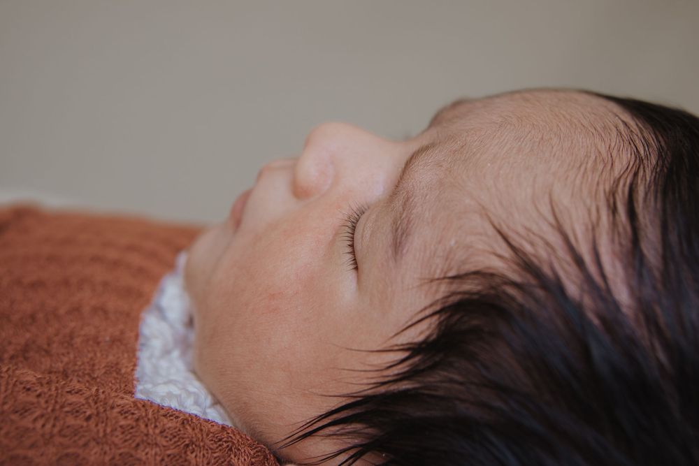 Peaceful newborn baby sleeping on a rust-colored blanket, with dark hair and delicate features in soft light.