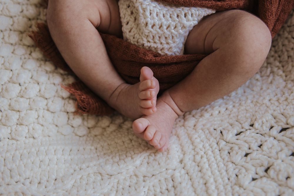 Close-up of a baby's chubby legs and tiny feet resting on a cream chunky knit blanket with a rust-colored wrap.