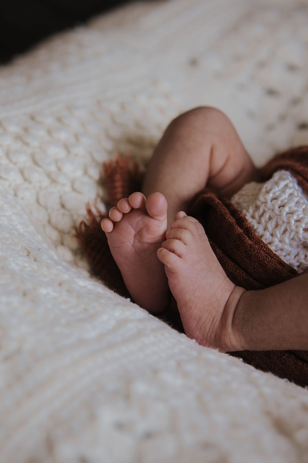 Close-up of tiny newborn baby feet resting on a soft cream knit blanket with a brown swaddle wrap during in- home photo session in Epping