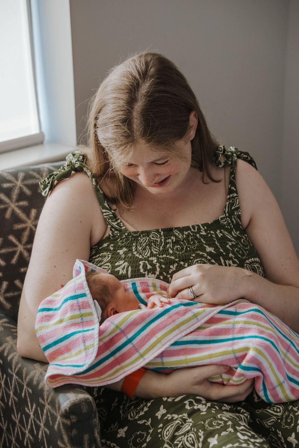 Mother in green dress tenderly holds newborn baby wrapped in colorful striped blanket while seated in patterned chair.