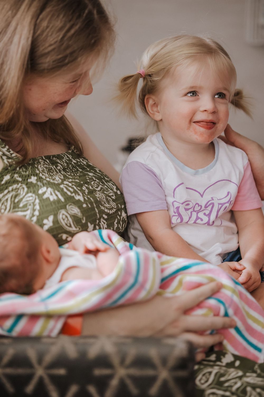 A smiling toddler girl in a sister shirt sits with her mother holding a newborn baby wrapped in a striped blanket.