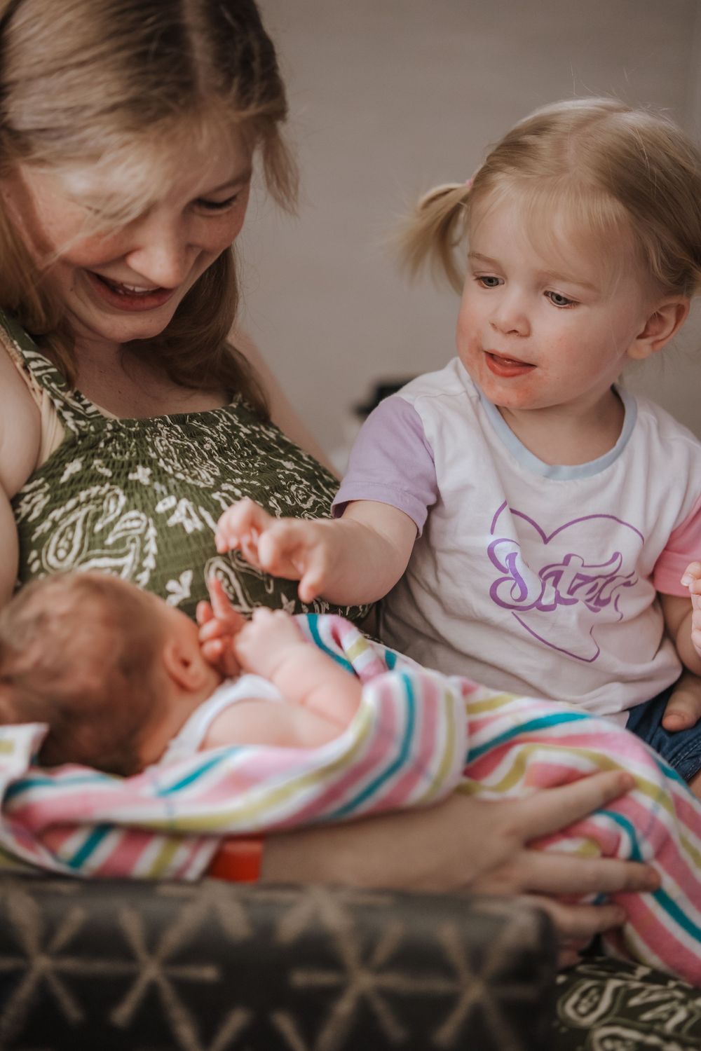 A smiling mother holds her newborn baby while a toddler girl in a Sister shirt reaches out to touch the infant.